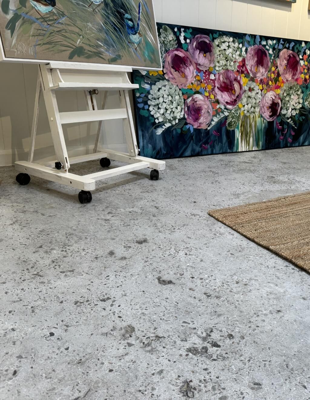 Concrete floor with a white easel holding a painting, and a large canvas with floral artwork leaning against the wall. A portion of a woven rug is visible on the right side.