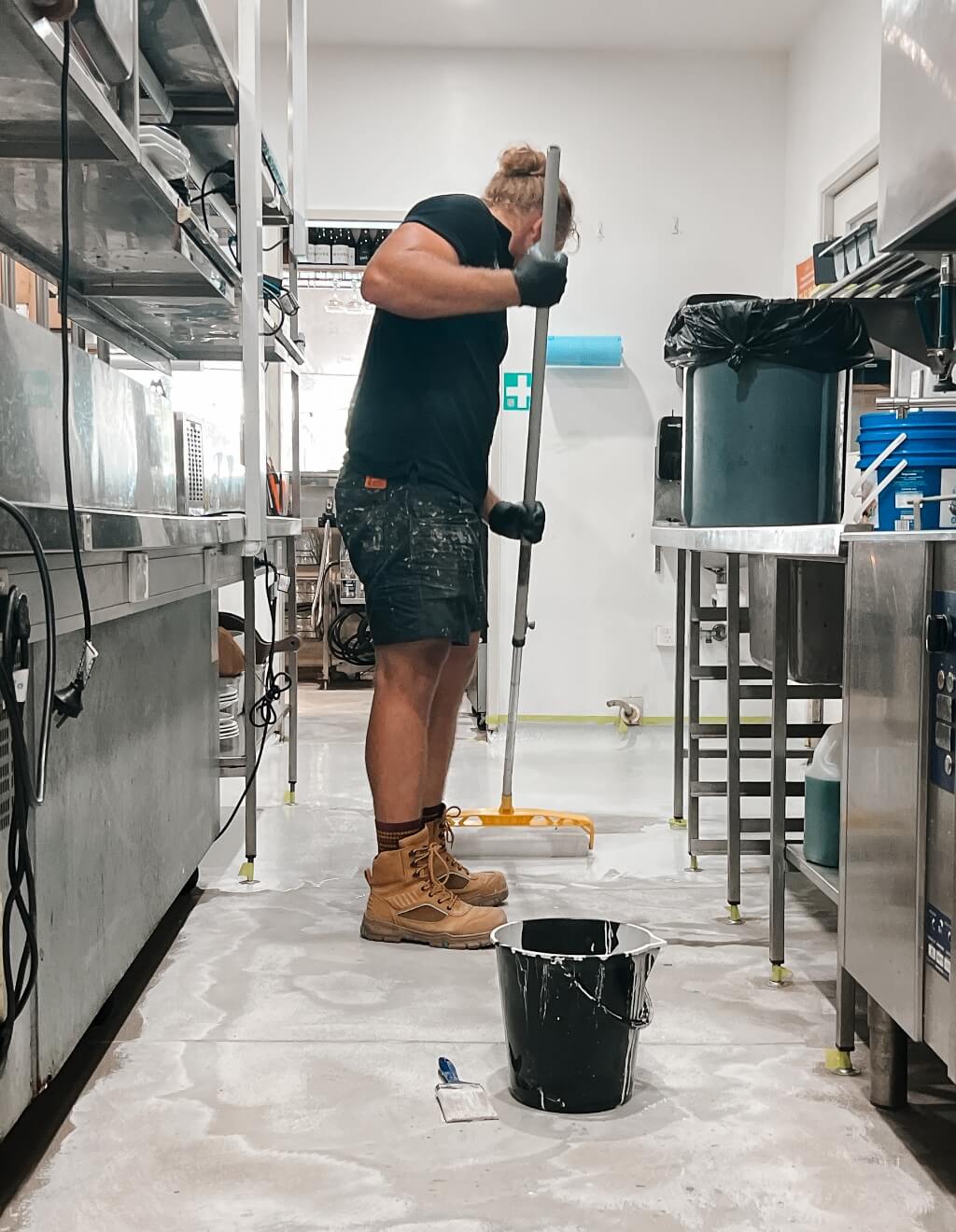 A person mops the floor in an industrial kitchen. They are wearing black gloves, a black shirt, and work boots. Cleaning supplies and equipment are visible around the area.