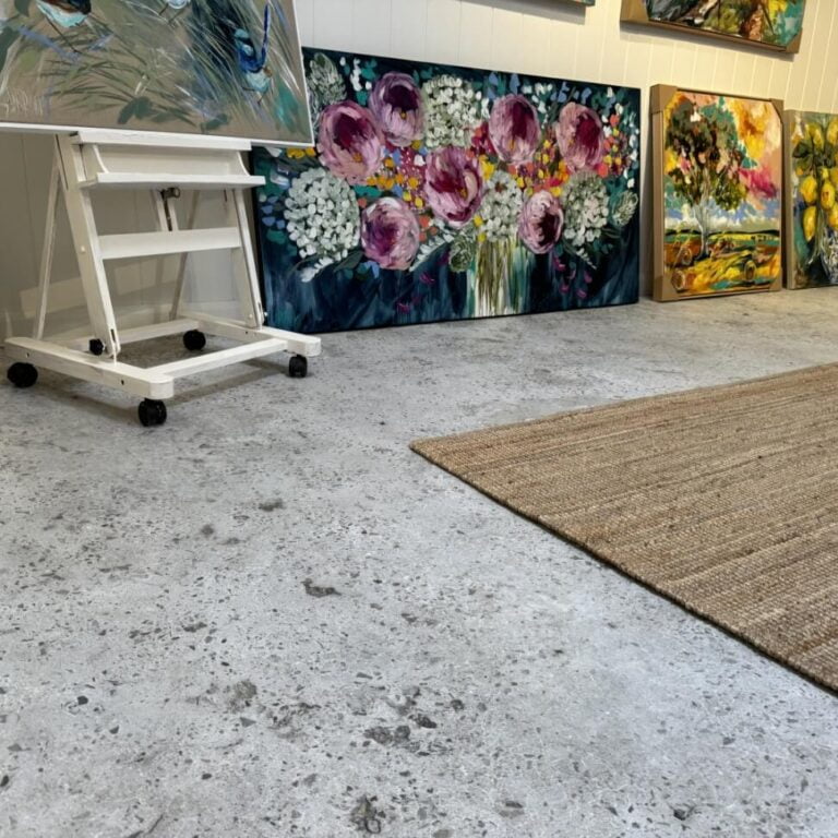 Art studio with various colorful paintings displayed on the wall and on an easel, featuring floral and landscape themes, and a textured concrete floor with a woven rug.