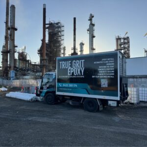 A truck with "True Grit Epoxy" branding is parked on a construction site with large industrial structures in the background. The truck's side displays contact information and services offered.