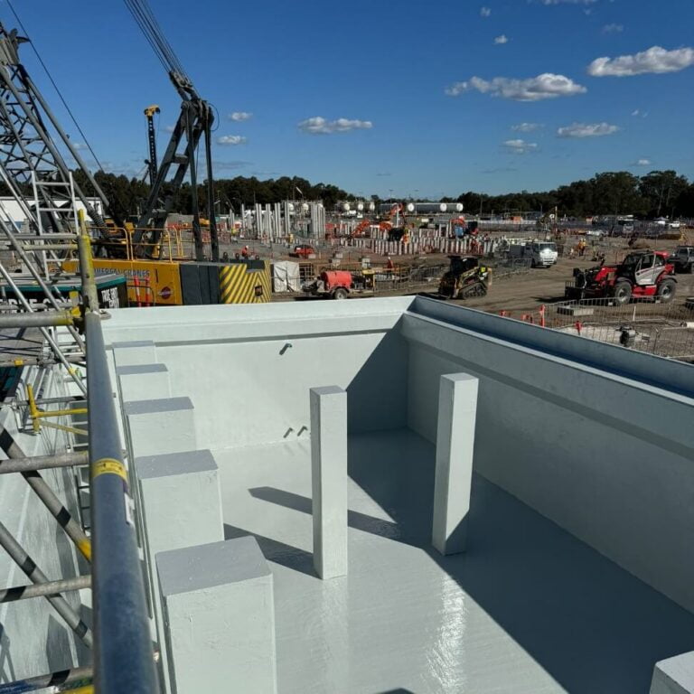 Construction site featuring heavy machinery and scaffolding under a blue sky. The foreground includes a newly completed concrete structure, with workers and additional construction activities in the background.