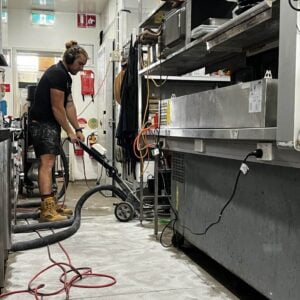 A person wearing headphones and work boots uses a floor cleaning machine in a commercial kitchen.