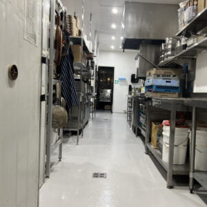 Commercial kitchen with stainless steel shelves and counters, various cooking utensils, and equipment. The floor is clean, and a door is visible at the far end of the room.