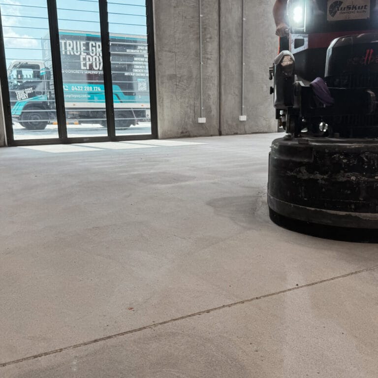 A floor polishing machine is in use inside a large room with concrete flooring. Sunlight comes through tall windows, and a truck is visible outside.