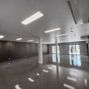 A spacious, empty room with polished concrete floors and overhead fluorescent lighting. Large windows on one side let in natural light.