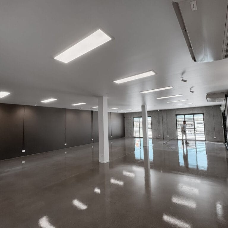 A spacious, empty room with polished concrete floors and overhead fluorescent lighting. Large windows on one side let in natural light.