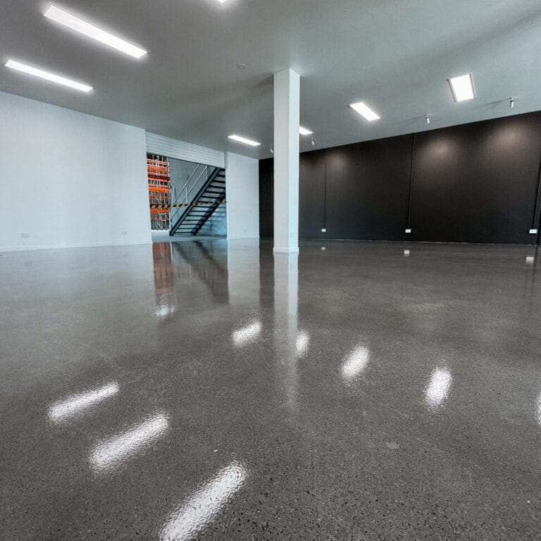 A spacious, empty room with polished concrete floors, white and black walls, ceiling lights, and a metal staircase leading to an upper level.