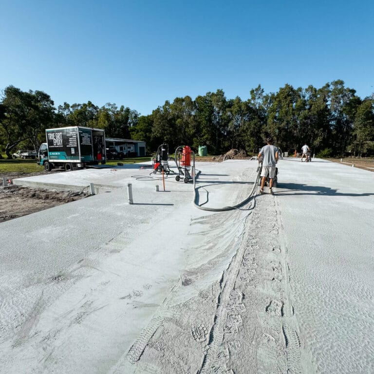 Workers are smoothing a large concrete slab on a construction site with machinery. A truck and trees are visible in the background.