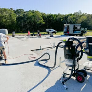 Workers operate machinery on a concrete surface at a construction site with a trailer, water tank, and forested area in the background.