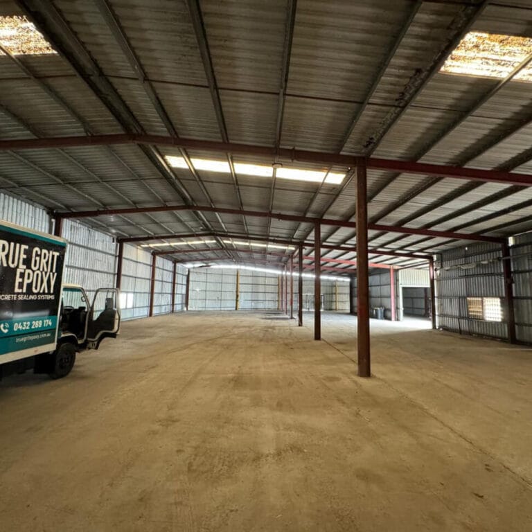 A large, empty warehouse interior with metal walls and ceiling beams. A truck with "True Grit Epoxy" branding is partially visible on the left.