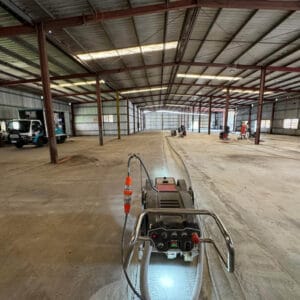 An empty warehouse with a concrete floor under construction, featuring machinery and two vehicles on the left side.