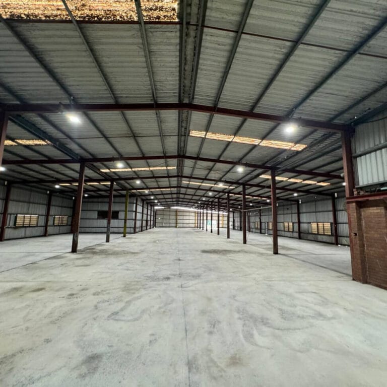 Spacious empty warehouse interior with metal beams, concrete floor, and corrugated roof, lit by overhead lights.