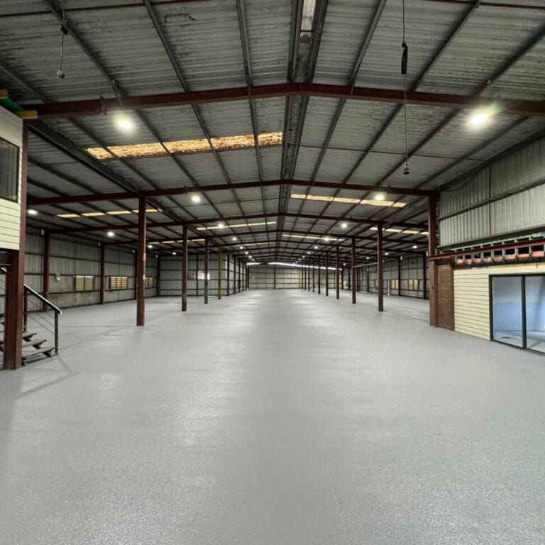 Large, empty warehouse interior with high ceilings, metal support beams, and concrete flooring. Natural light filters through skylights.