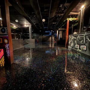 Dimly lit room with a glittering, reflective black floor, graffiti-covered walls, and various light fixtures, creating a vibrant atmosphere.