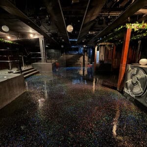 A dimly lit nightclub features a floor covered in multicolored glitter, with overhead lights and lush greenery on one side.