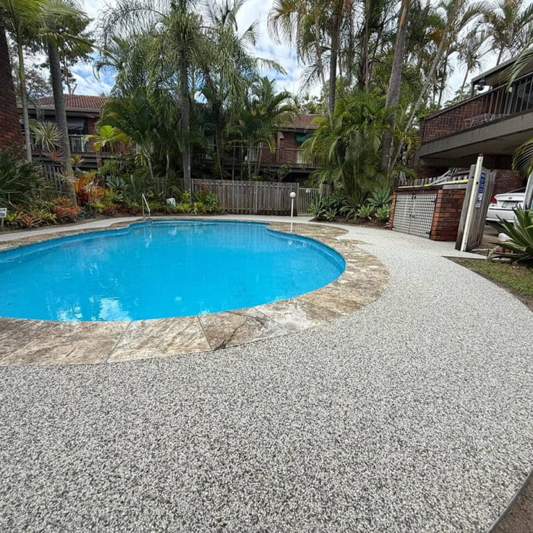 A small blue swimming pool with a speckled concrete surround, bordered by tropical plants and trees, situated in a yard with a wooden fence and brick buildings in the background.