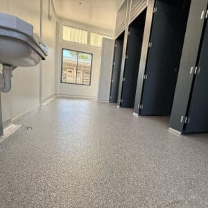 A public restroom with a gray textured floor, black stall doors, a wall-mounted sink, and a window letting in natural light.