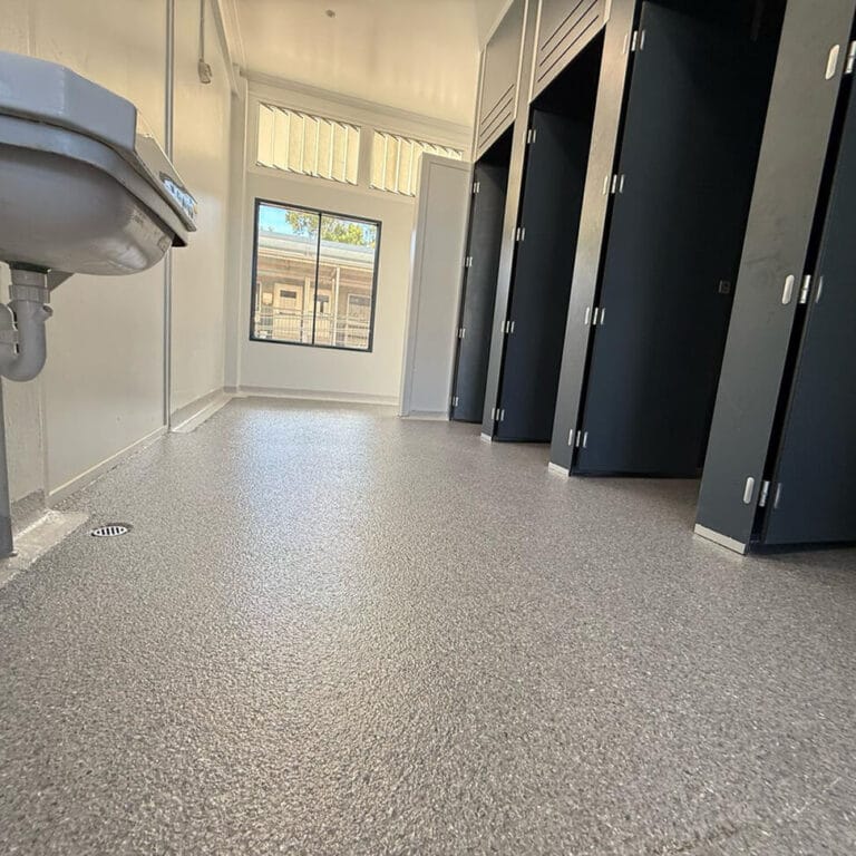 A public restroom with a gray textured floor, black stall doors, a wall-mounted sink, and a window letting in natural light.
