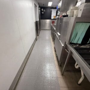 A clean commercial kitchen hallway with dishwashing equipment, stacked plates, utensils, and gray tiled flooring.