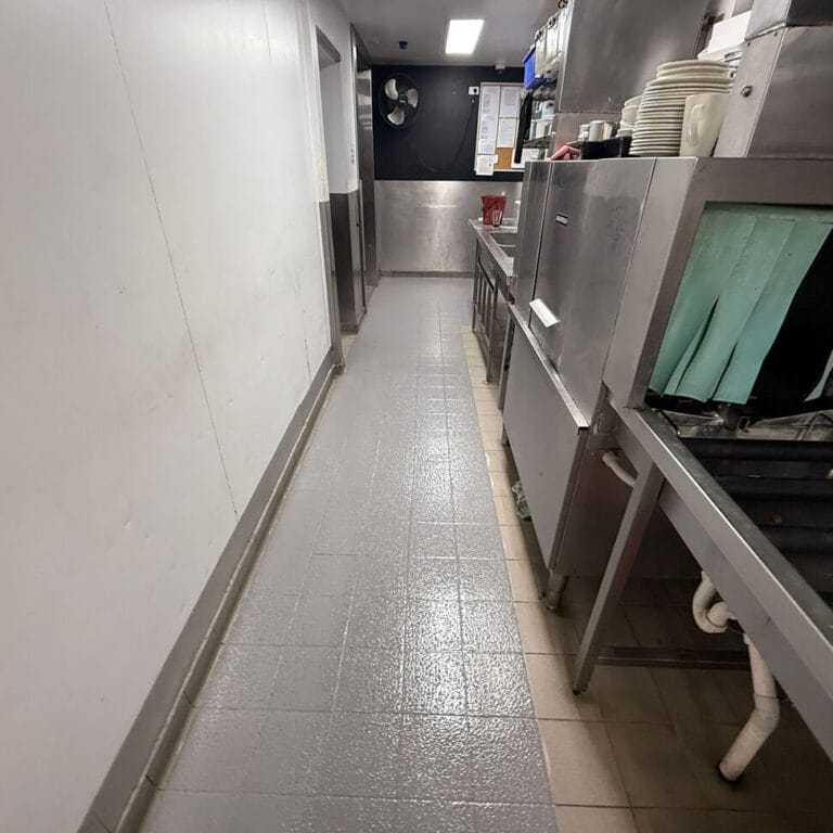 A clean commercial kitchen hallway with dishwashing equipment, stacked plates, utensils, and gray tiled flooring.