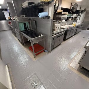 A clean commercial kitchen with stainless steel appliances, empty countertops, shelves with dishes, and a shiny tile floor.