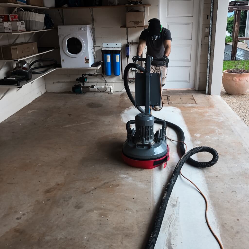 Person using a floor scrubber to polish the concrete floor in a garage with shelves, a washing machine, and a water filtration system in the background.