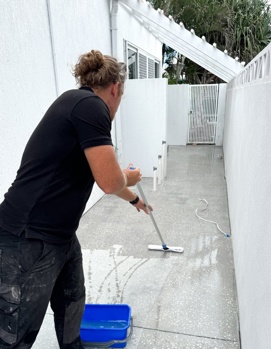 A person with long hair in a bun is cleaning a narrow outdoor pathway using a mop and a blue bucket, likely preparing the area for concrete resurfacing. The walls are white and appear freshly painted.