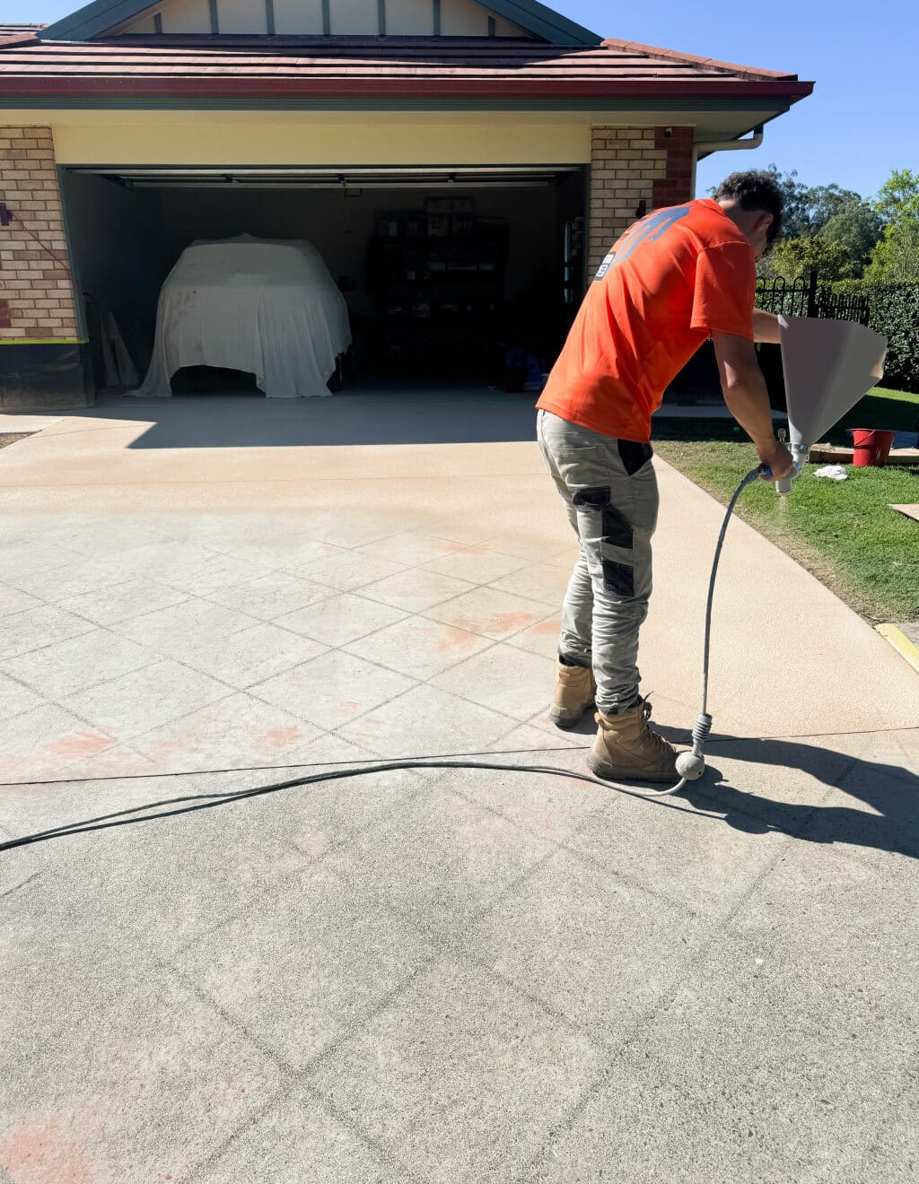 A person in a red shirt and work boots uses a spray hose on a driveway in front of an open garage.