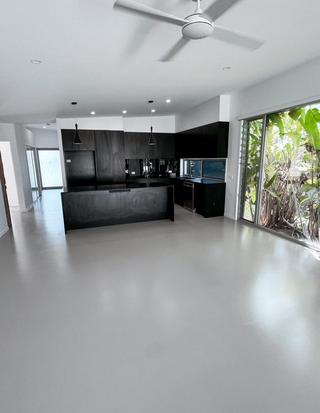 Modern kitchen with dark cabinetry, pendant lights, and large windows overlooking greenery. White walls and a ceiling fan complement the light gray epoxy flooring, adding a sleek touch to the space.