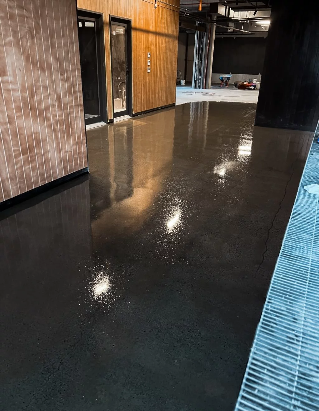 A polished concrete floor reflects light in a modern indoor space with wooden and black walls, showcasing the excellence of concrete polishing. Construction equipment is visible in the background.