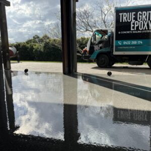 Epoxy-coated floor reflects light in a garage with open doors. Outside, a truck with "True Grit Epoxy" branding is parked.