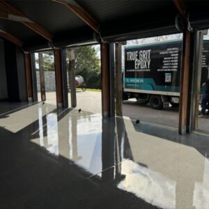 A polished concrete floor in a large garage with open doors. Outside, a truck with "True Grit Epoxy" branding is parked, and a person is seen partially entering or exiting the garage.