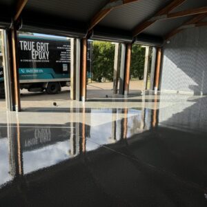 A polished concrete garage floor reflects light, with a truck featuring "True Grit Epoxy" parked outside under an open garage door.