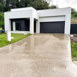 A modern, single-story house with white walls, a black garage door, and a neatly manicured lawn. The driveway is paved and slightly wet. House number 70 is visible on a post beside the driveway.