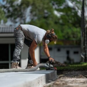 Person wearing construction gear and ear protection uses a power tool on a concrete surface outdoors. Trees and a building are visible in the background.