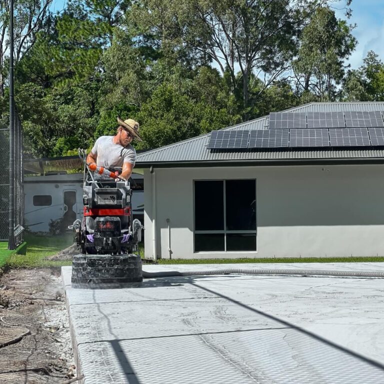 A person operates a power trowel on a concrete surface outside a building with solar panels on the roof. Trees are visible in the background.