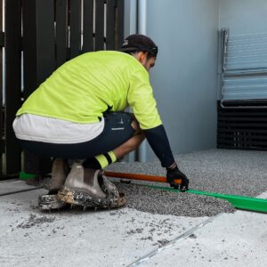 A person in protective gear and a neon shirt is spreading a mixture over a surface using a tool, near a gate and some stacked chairs.