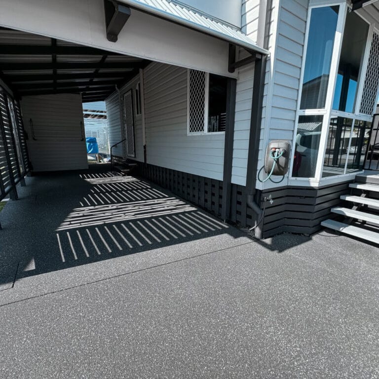 Covered parking area beside a light gray house with a windowed corner and side steps. The ground has a textured gray surface.