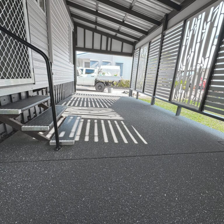 Covered outdoor area with patterned sunlight on a speckled floor, metal steps, and slatted privacy screens. White truck visible outside near a neighboring house.