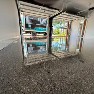 Shiny epoxy-coated garage floor reflecting light, with a truck parked outside displaying "True Grit Epoxy" branding.