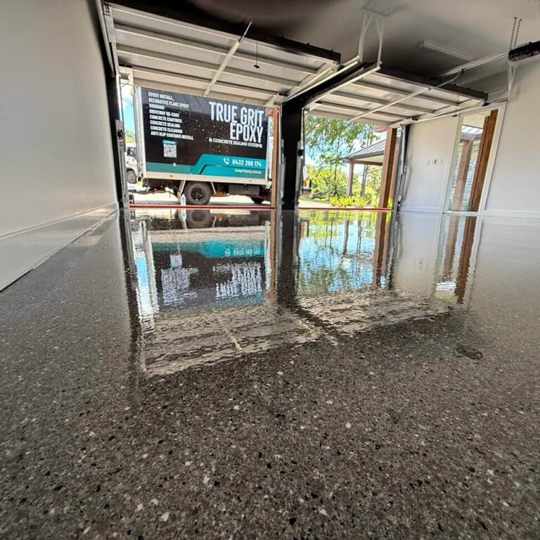 Shiny epoxy-coated garage floor reflecting light, with a truck parked outside displaying "True Grit Epoxy" branding.