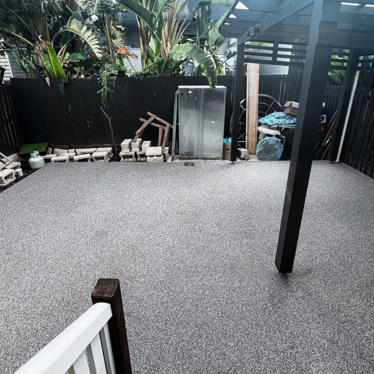 A newly laid concrete patio with black pergola beams and scattered construction materials, bordered by a black fence and tropical plants.