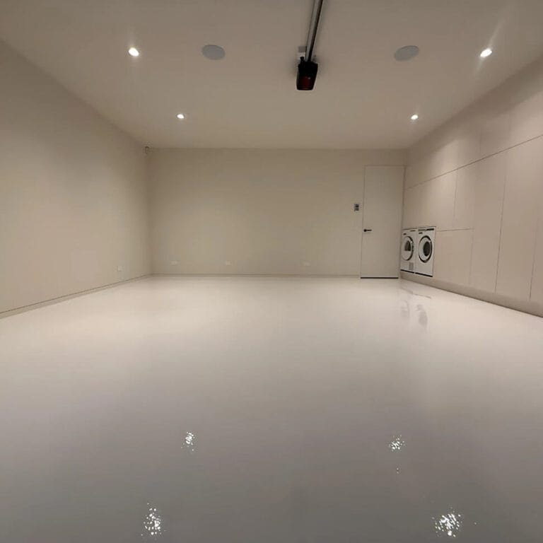 Empty, clean garage with a polished white floor, beige walls, and ceiling lights. A washer and dryer are installed in the back corner near a closed door.