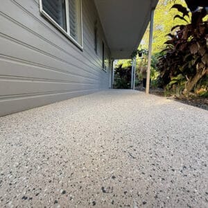 A textured, speckled concrete porch runs alongside a light grey house with siding, bordered by plants and a covered roof area.