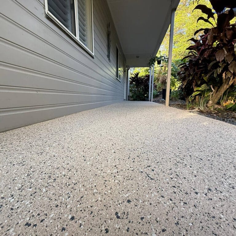 A textured, speckled concrete porch runs alongside a light grey house with siding, bordered by plants and a covered roof area.
