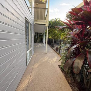 A concrete walkway runs alongside a modern building with white siding, bordered by a metal fence and tropical plants.
