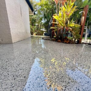 A polished concrete patio reflects surrounding plants and a fence in an outdoor garden area near a light-colored brick wall.