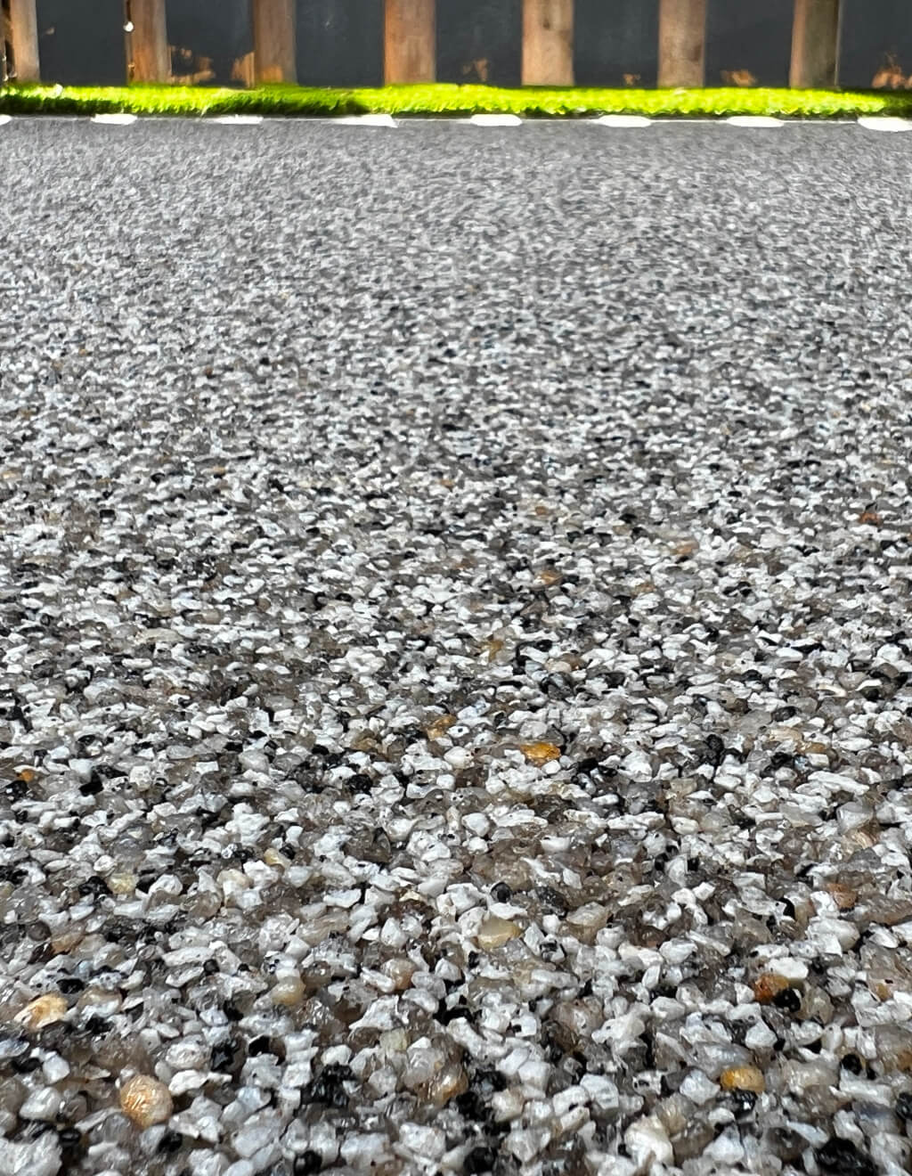 Close-up of a textured gravel surface with small pebbles in various shades of gray and black. Green plants and a wooden fence are blurred in the background.