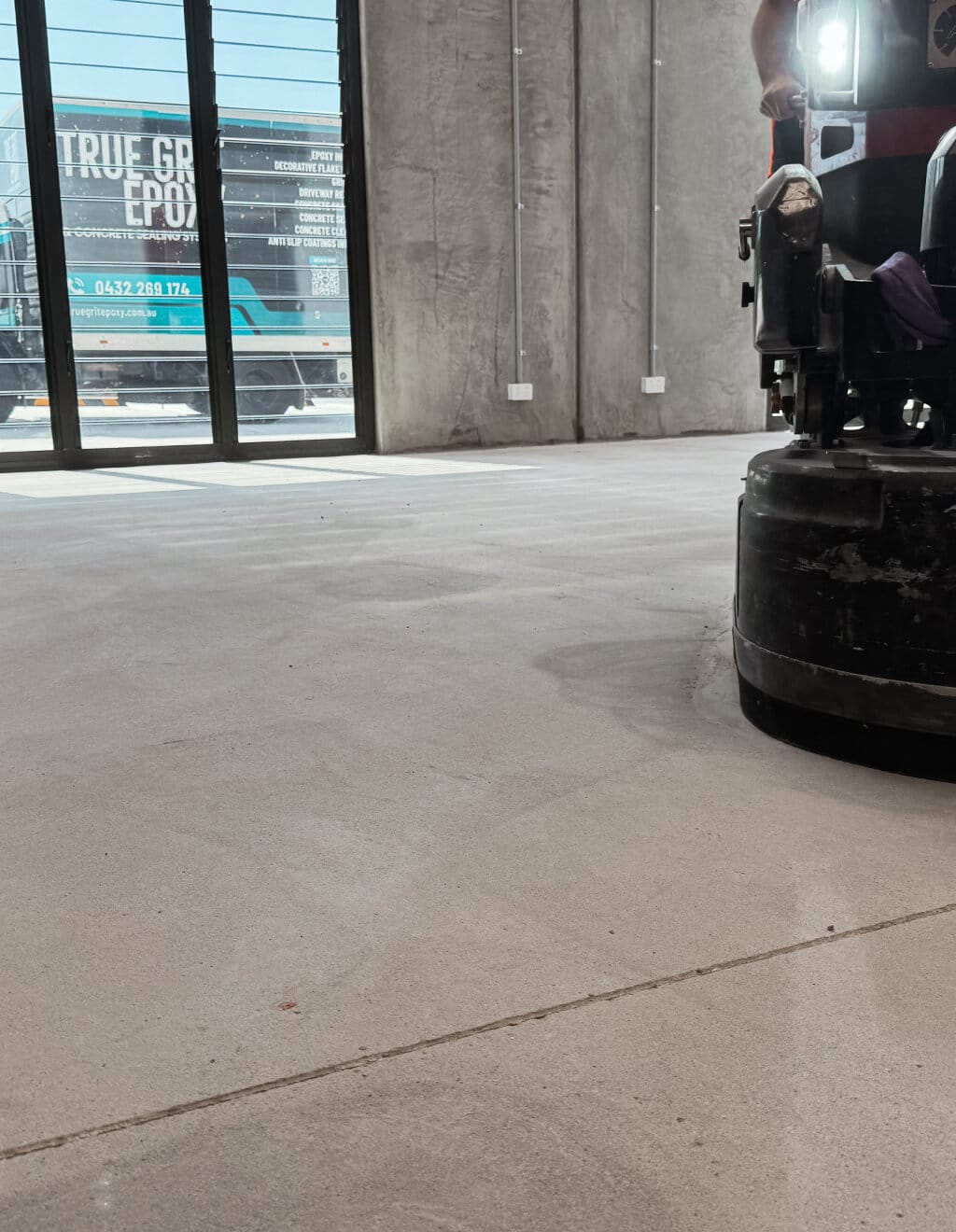 A close-up of a floor polishing machine in action on a concrete floor inside a building with large windows. A truck is visible outside in the background.